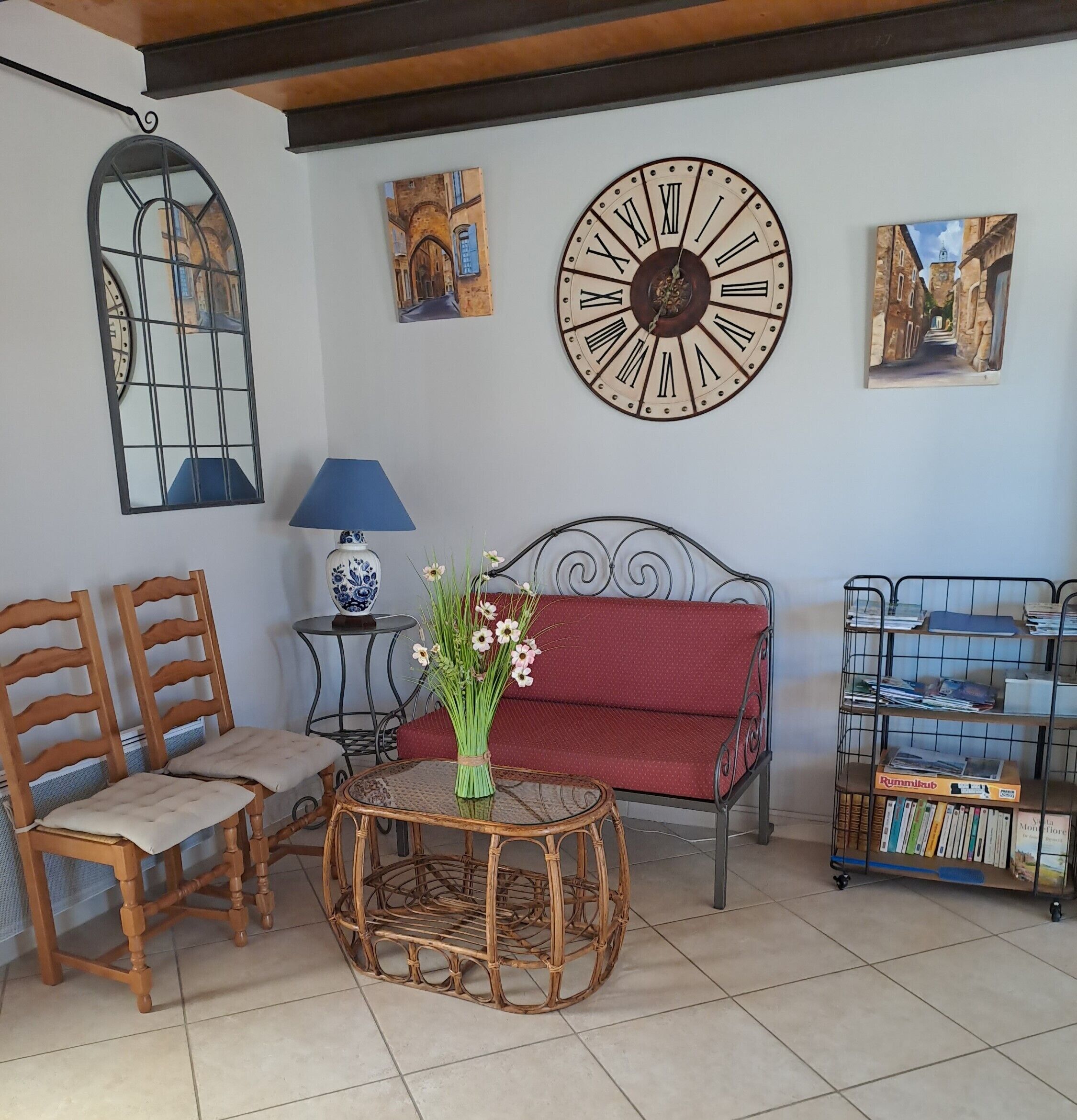 A living room with a red sofa, two wooden chairs, a round wicker table with a vase of flowers, a wall clock, and a bookshelf.