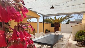 An outdoor patio with a table and chairs, a covered area with a white umbrella, and a yellow building in the background.
