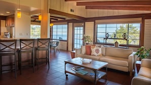 A spacious living area with a large window, a sofa, a coffee table, and a kitchen area in the background.