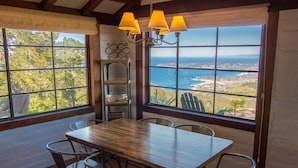 A dining area with a wooden table and metal chairs, large windows with a view of the ocean and trees, and a chandelier with multiple lamps.