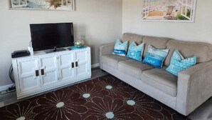 Una sala de estar con una televisión de pantalla plana sobre un mueble blanco, un sofá beige con cojines azules y una alfombra marrón estampada.
