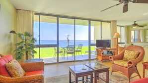 Uma sala de estar espaçosa com uma grande porta de vidro deslizante que dá para uma varanda com vista para o oceano. Há uma televisão, uma mesa de centro e móveis de vime.