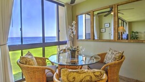 Quarto com mesa de vidro, cadeiras de vime, ventilador de teto e grande janela com vista para o mar.