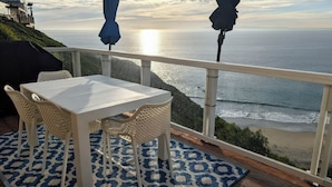 Ein Balkon mit weißem Tisch und Korbstühlen, mit Blick auf einen Strand und den Ozean.