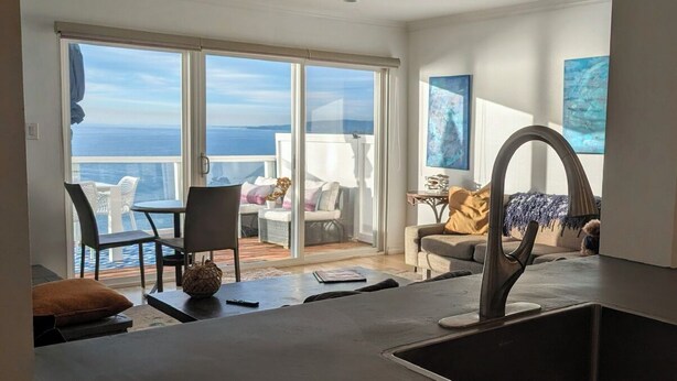 Ein modernes Wohnzimmer mit einem großen Schiebeglastür, die zu einem Balkon mit Meerblick führt. Es gibt eine Essecke mit Tisch und Stühlen sowie eine Couch mit einer dekorativen Vase.