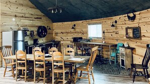 Interior rústico de madera con techo alto, una mesa de comedor rodeada de sillas y una zona de cocina con electrodomésticos.