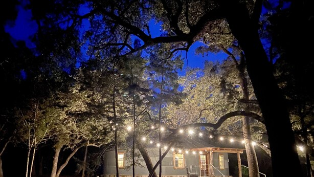 Una cabaña rústica con luces colgantes, rodeada de árboles, y una mesa de picnic en un área boscosa.