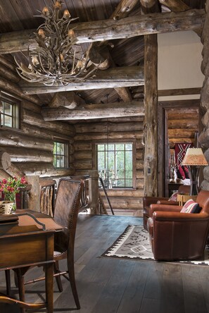 A rustic log cabin interior with a chandelier, wooden furniture, and a large window.