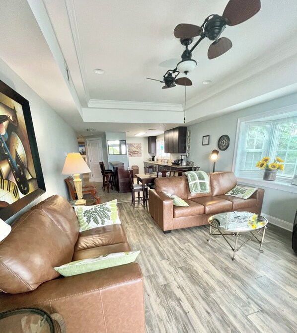 A spacious living room with a brown leather sofa, a glass coffee table, and a ceiling fan.