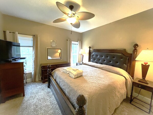 A hotel room with a bed, a television, a ceiling fan, a nightstand with a lamp, and a dresser.