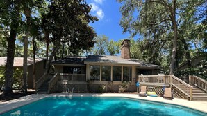 Un área de piscina con terraza de madera, zona de estar cubierta y una casa con chimenea.