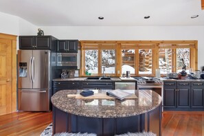 A modern kitchen with dark cabinetry, stainless steel appliances, a granite countertop island, and a view of a snowy landscape through large windows.