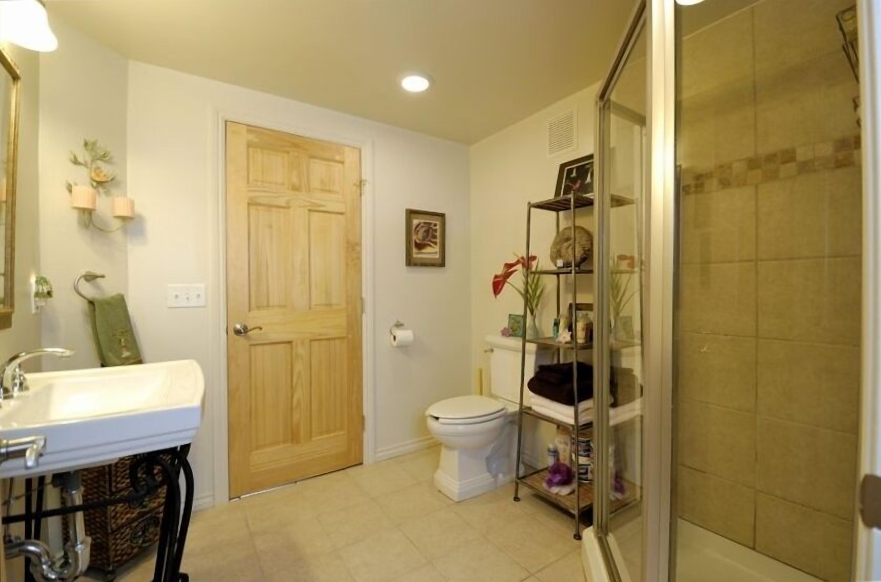A bathroom with a white sink, toilet, and shower. There is a wooden door and a shelf with toiletries.