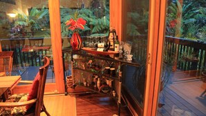 A wooden deck with a glass-enclosed dining area, a chair, and a shelf with wine bottles and glasses. - ~Peace and Tranquility in rain-forested Volcano on luxurious property ~ (Volcano)