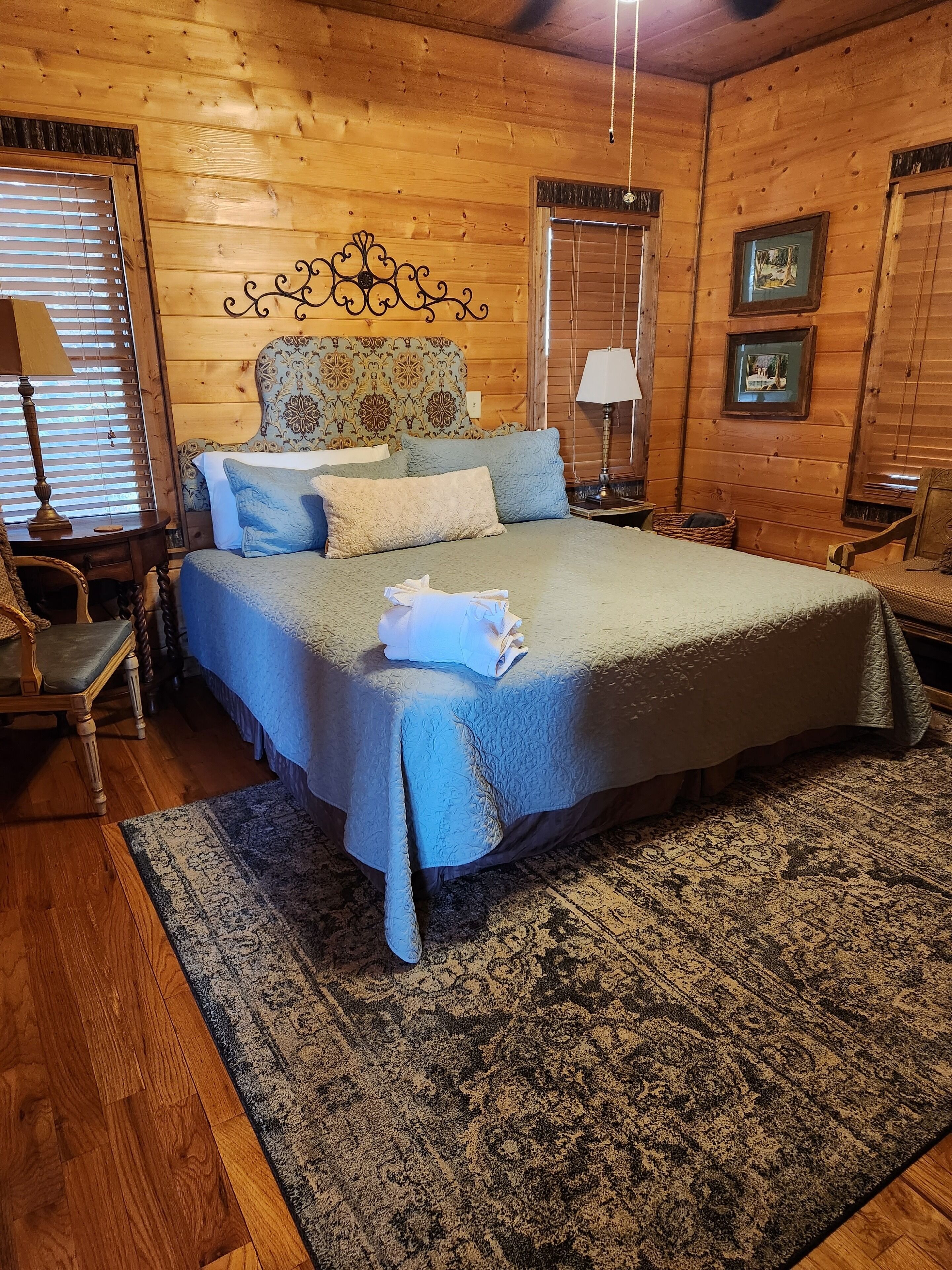 A wooden cabin bedroom with a large bed, a headboard with a floral pattern, a wooden floor, and a window with blinds.