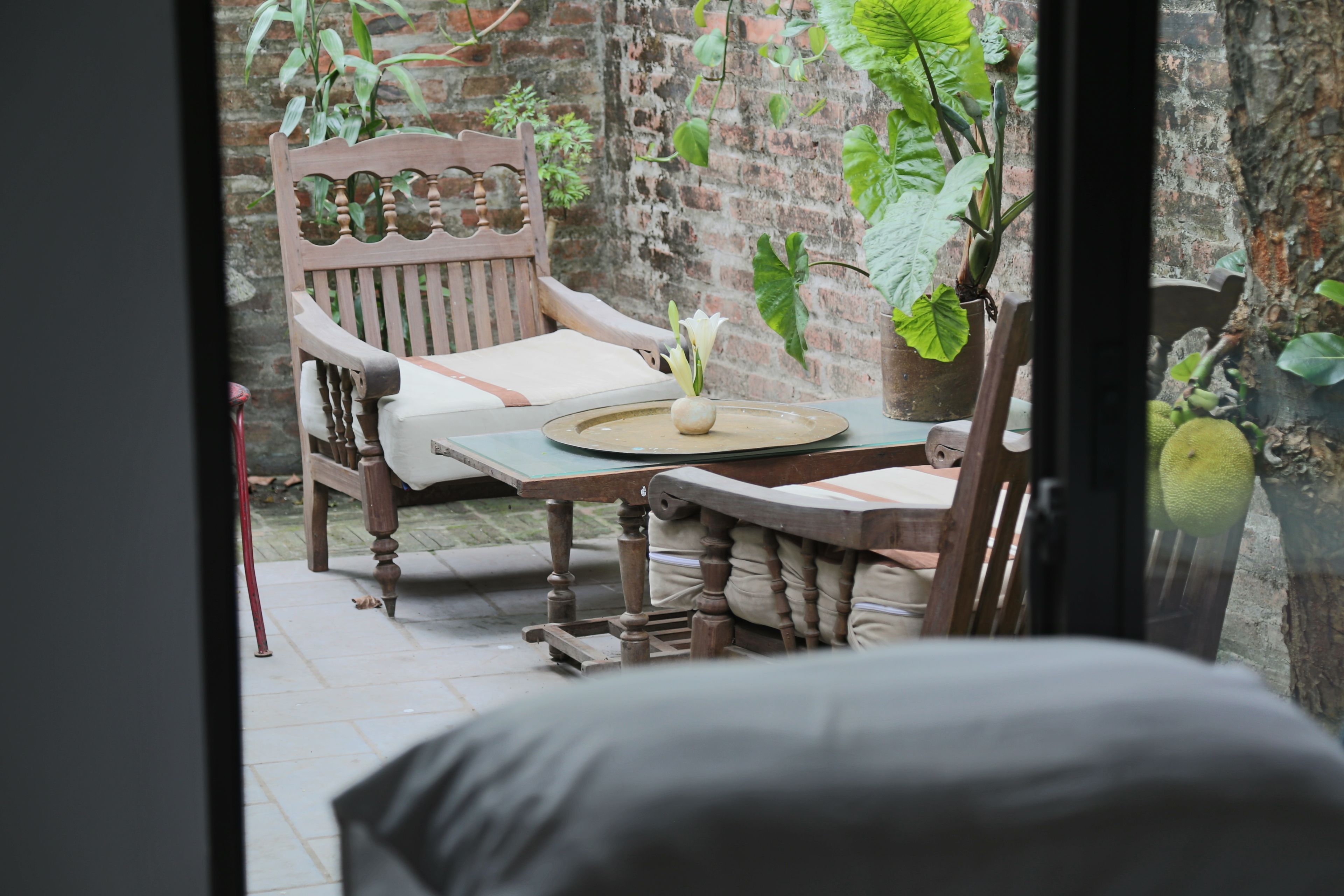 A wooden chair with a cushion, a small table with a vase, and a brick wall with plants.
