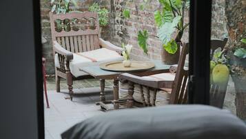 A wooden chair with a cushion, a small table with a vase, and a brick wall with plants.