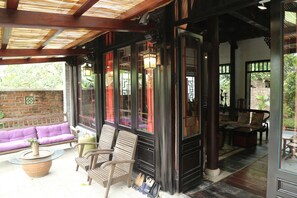 A covered outdoor seating area with wooden furniture, a glass-top table, and a brick wall. - Maison Haily (Hanoi)