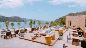 A terrace with lounge chairs and tables overlooking a body of water, with a stone wall and palm trees in the background.