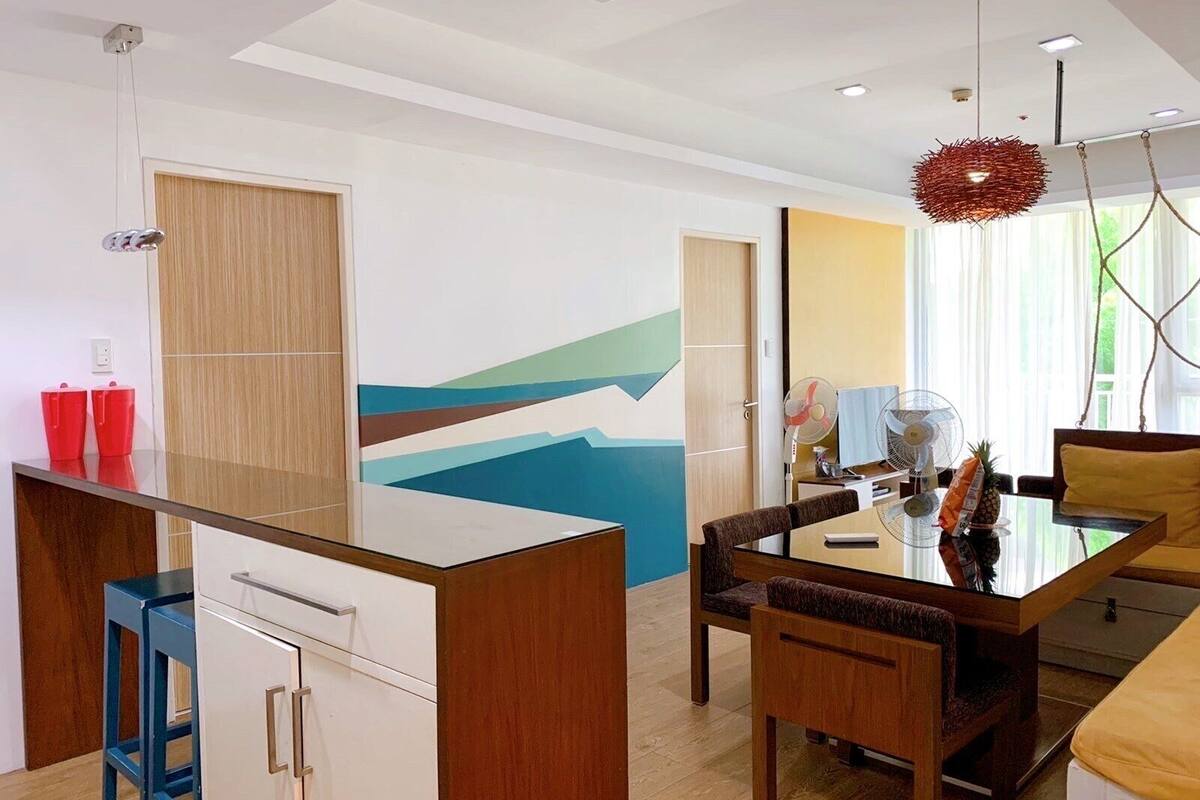 A modern living room with a dining area, a wooden cabinet, a glass table, and a colorful abstract wall.