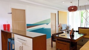 A modern living room with a dining area, a wooden cabinet, a glass table, and a colorful abstract wall.