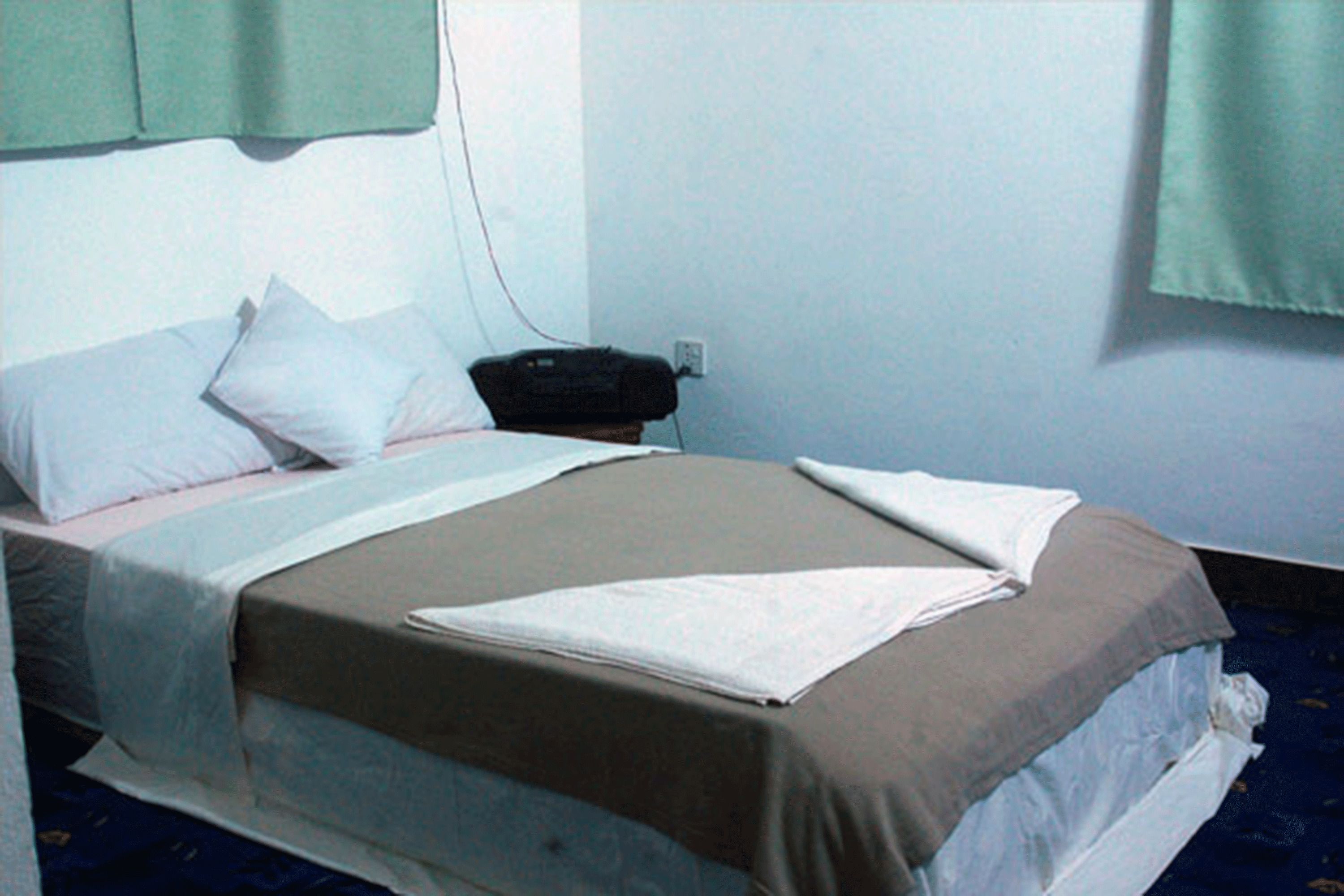 A single bed with a brown bedspread and white pillows, a bedside telephone, and a window with a green curtain.