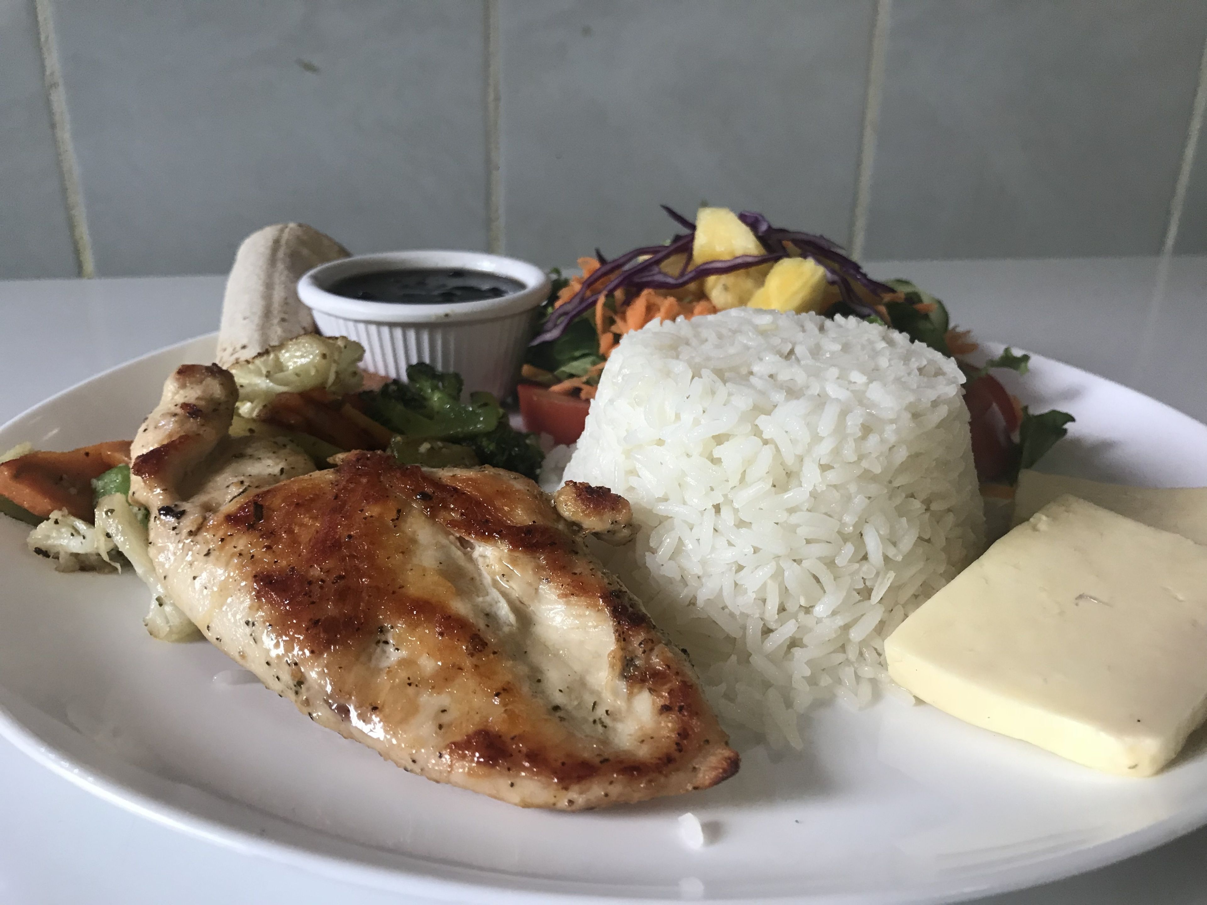 A plate with grilled chicken, white rice, a side salad, a small bowl of sauce, and a piece of butter.