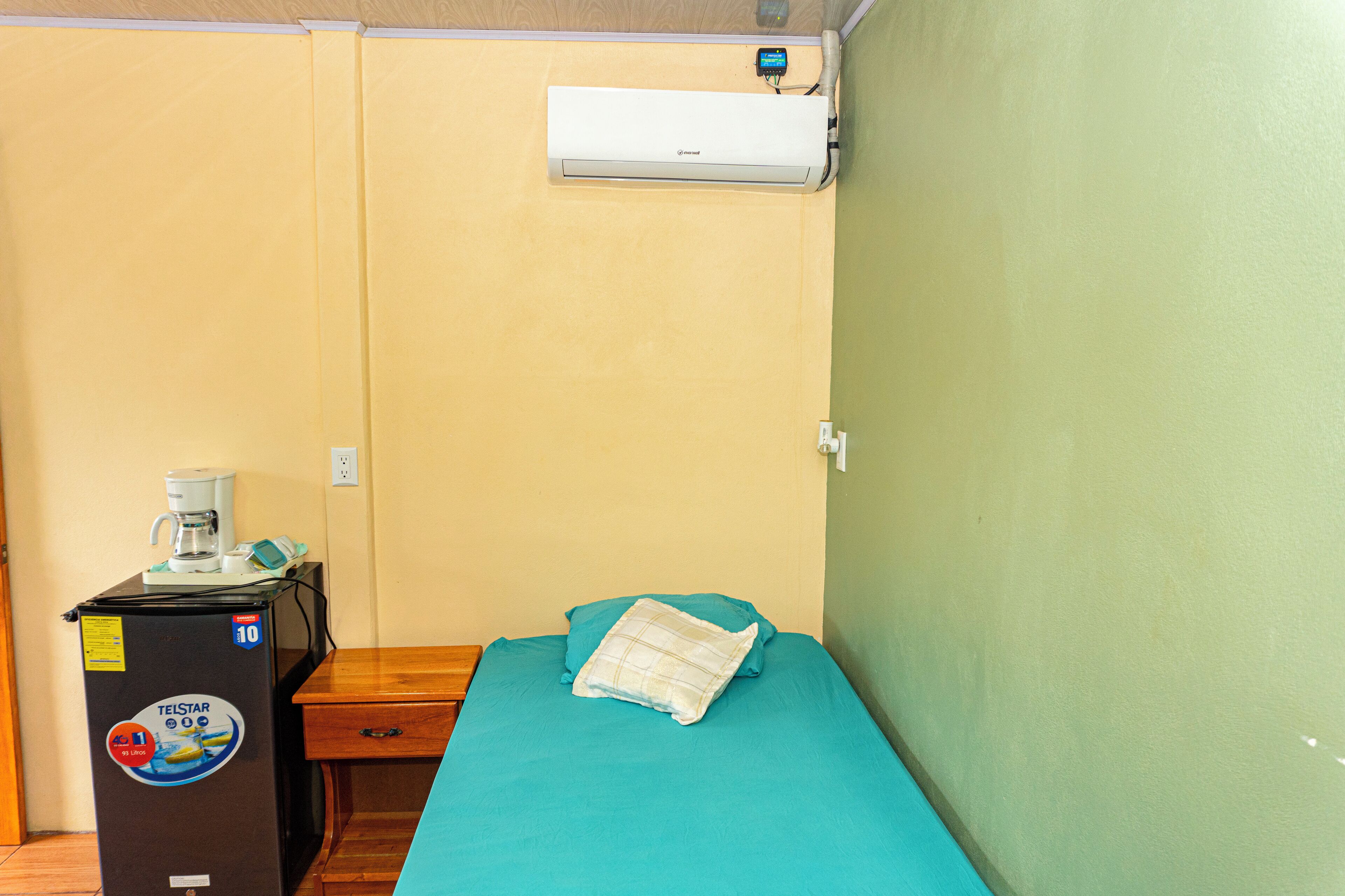 A small room with a bed, a desk with a coffee maker, and an air conditioner mounted on the wall.
