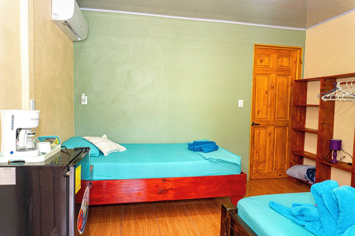 A small room with a bed, a wooden wardrobe, a desk with a coffee machine, and a wall-mounted heater.