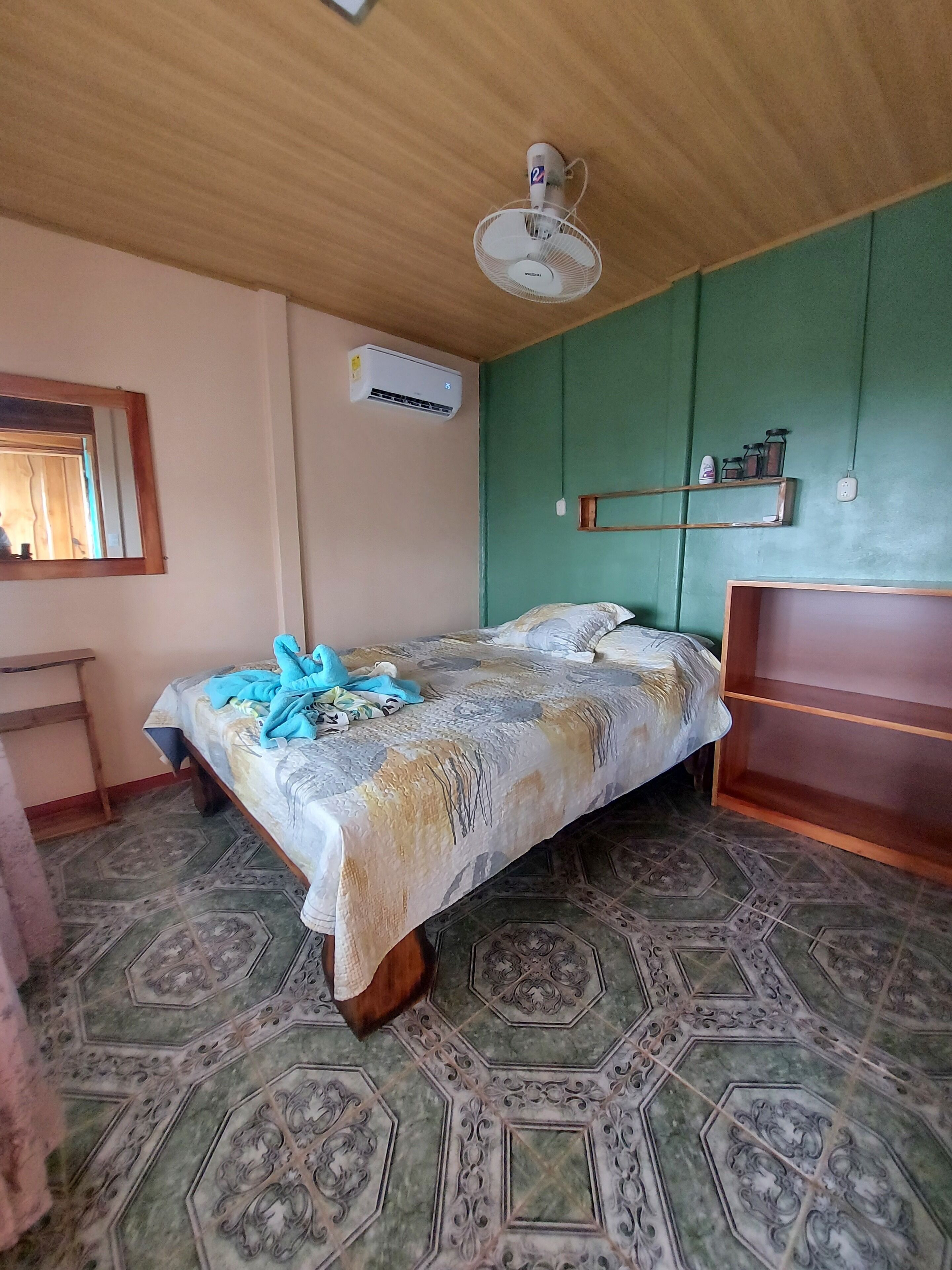 Une chambre comprenant un lit, une tête de lit en bois, un climatiseur fixé au mur, un miroir et une étagère avec divers objets.