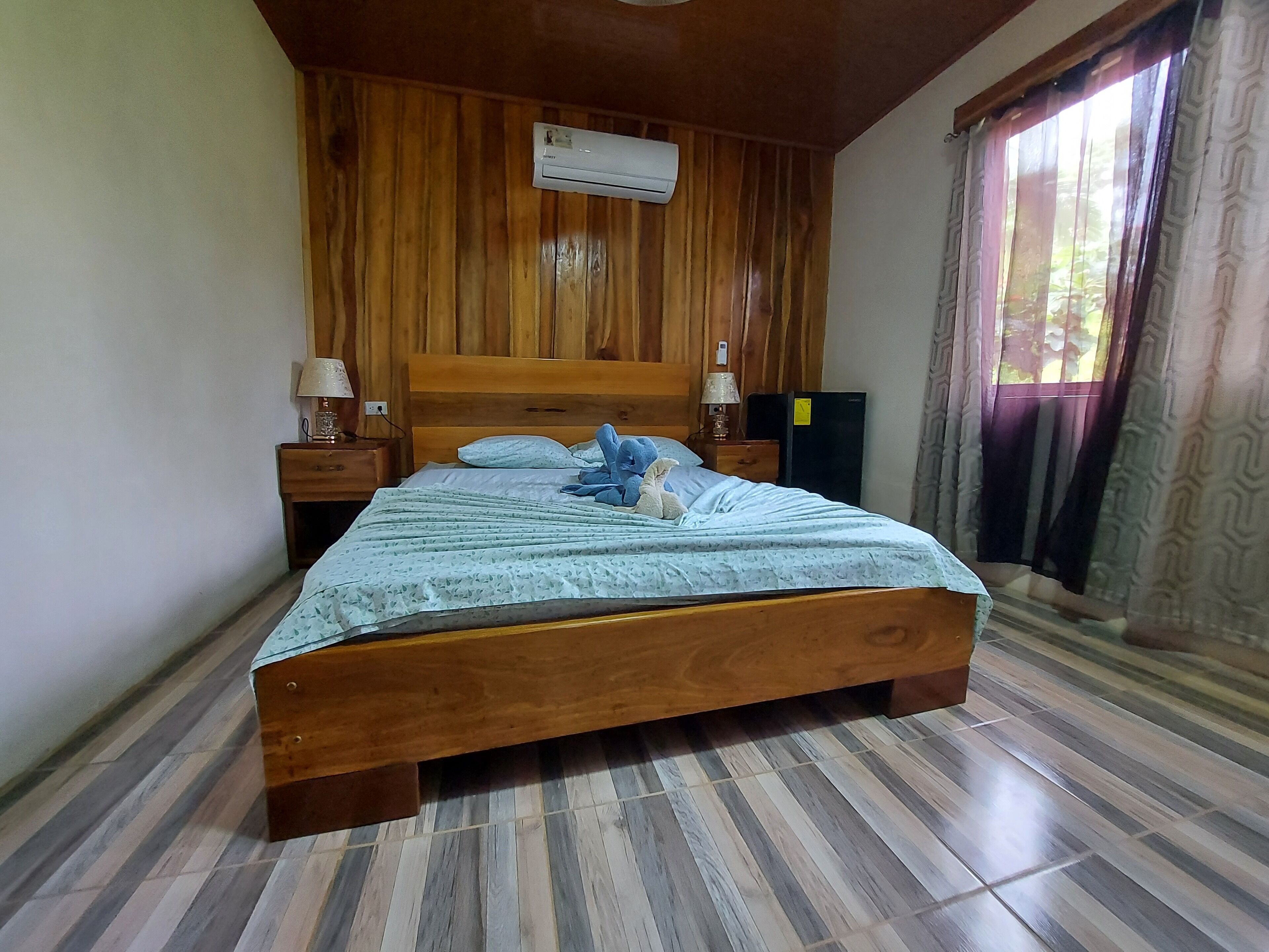 A wooden-paneled bedroom with a bed, two bedside tables, an air conditioner, and a window with curtains.