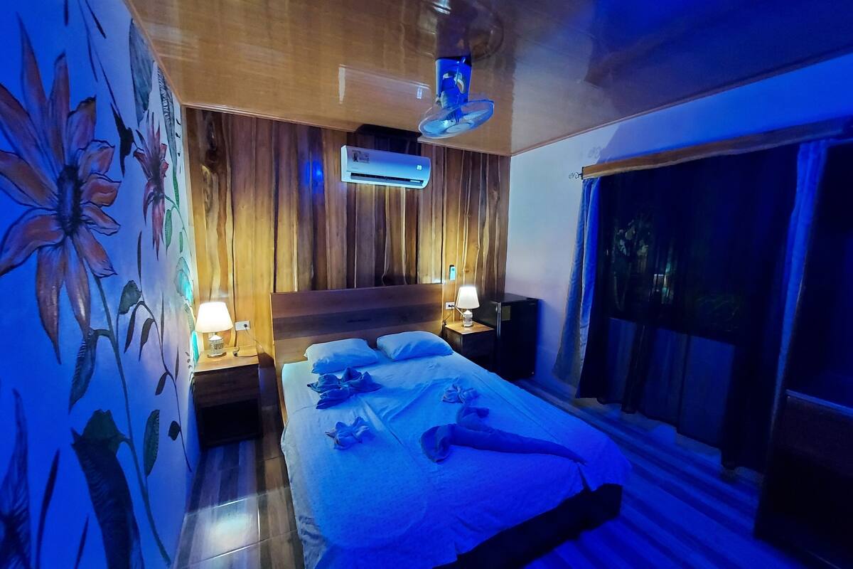 A bedroom with a wooden headboard, a bed with white linens, a nightstand with a lamp, and a wall-mounted air conditioner.