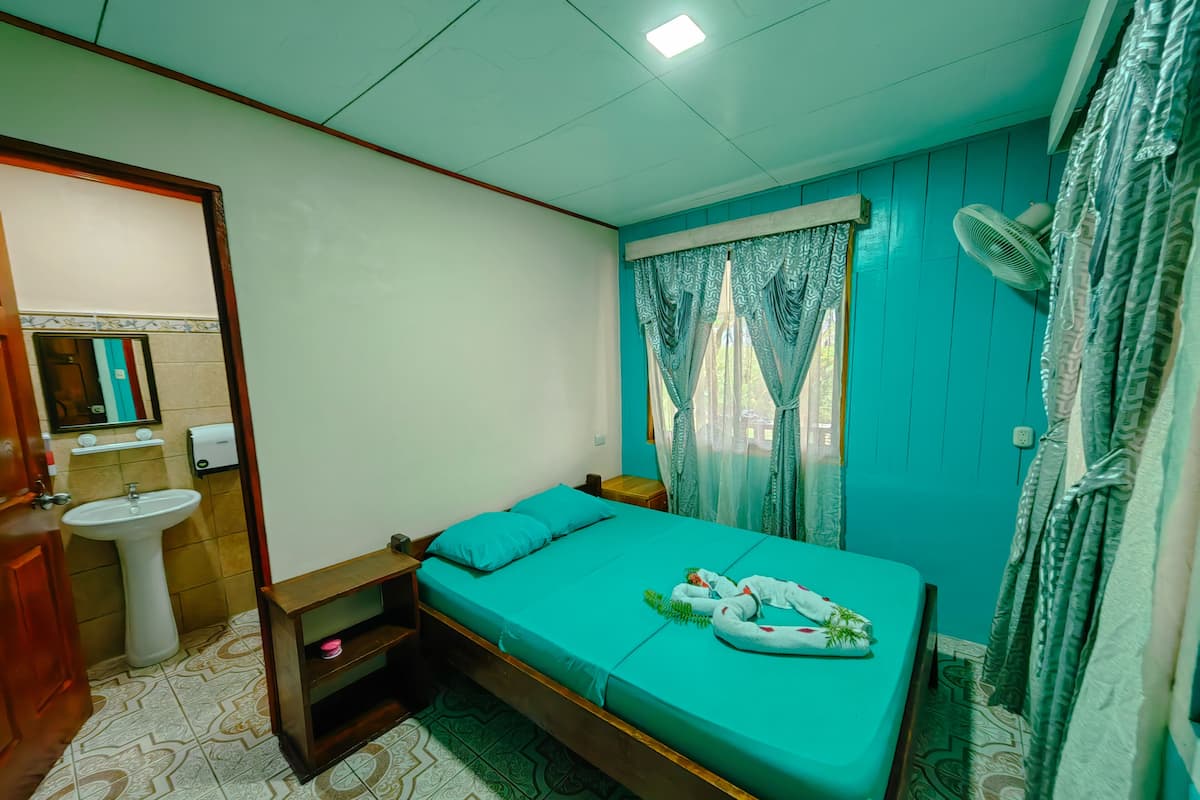 A bedroom with a bed, a bedside table, a window with curtains, a wall-mounted fan, and a bathroom with a sink and mirror.