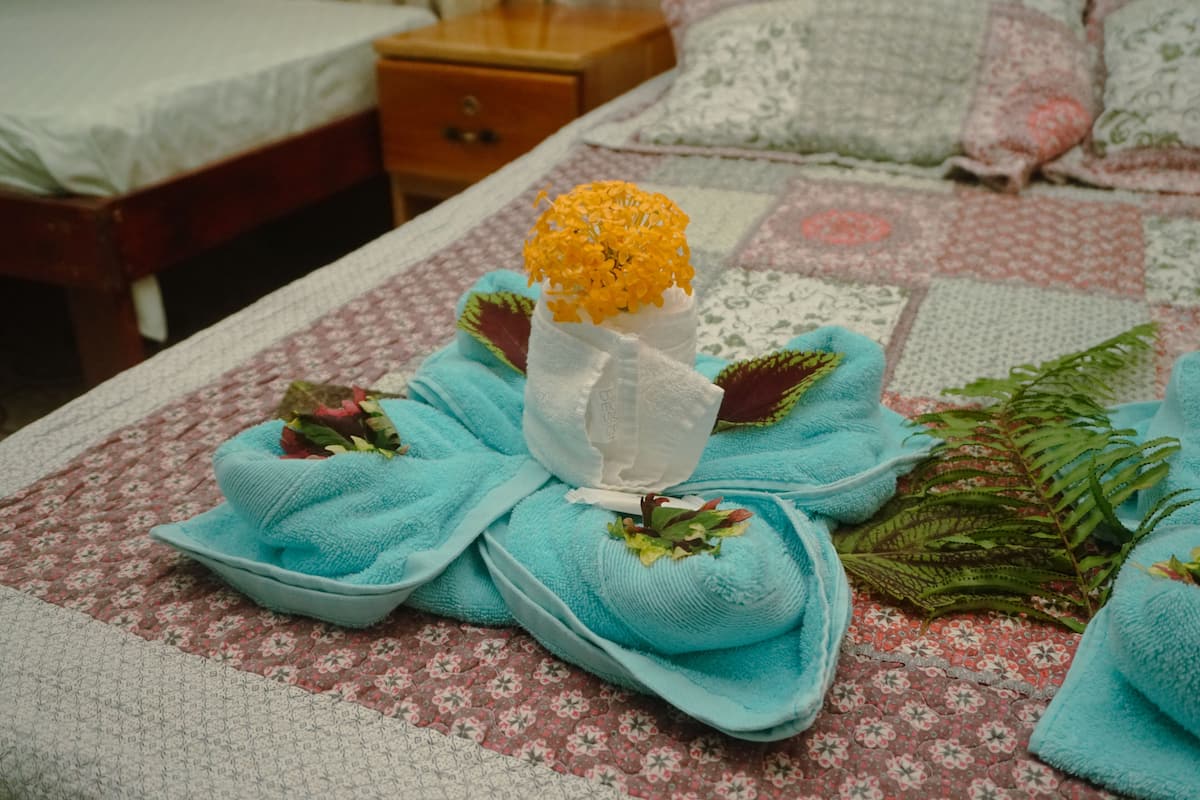 A neatly made bed with a floral-patterned bedspread, a vase with yellow flowers, and a blue towel with a leaf.