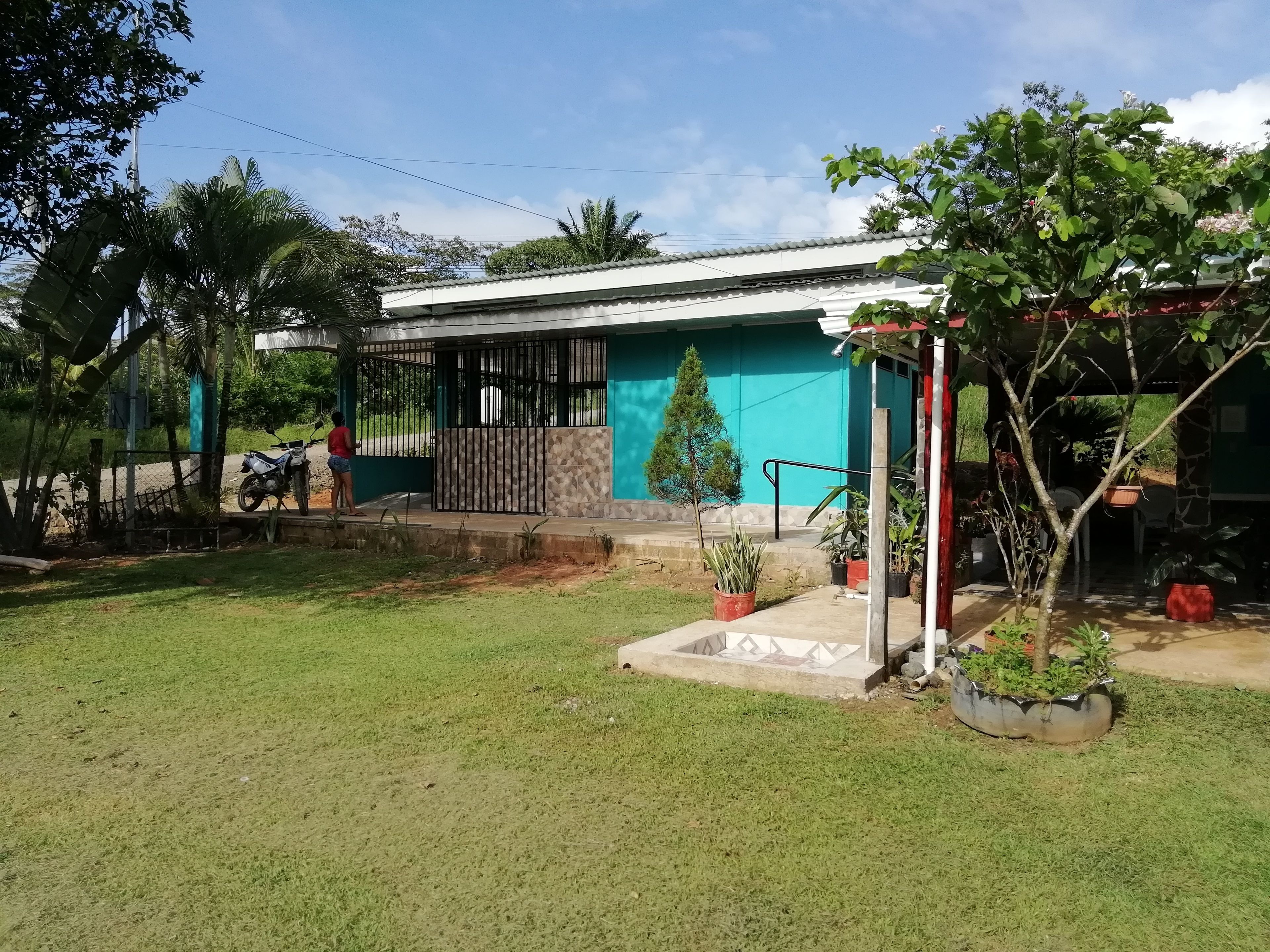 A single-story house with a white roof and teal walls, surrounded by a well-maintained lawn and potted plants.