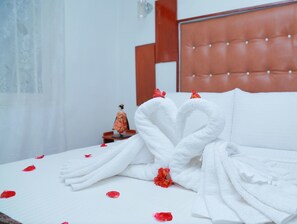 A neatly made bed with white towels arranged in a heart shape, red flower petals scattered on the bed, and a small figurine on the nightstand. - Canvi Villa (Nuwara Eliya)