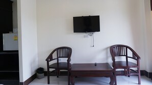 A room with a flat-screen TV mounted on the wall, two wooden chairs, a small table, and a refrigerator in the corner.