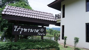 A sign with Thai script, a building with a tiled roof, and a garden area with small trees and shrubs.