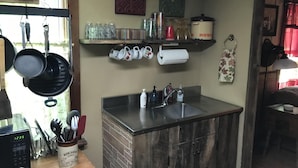 Een kleine keuken met een spoelbak, een magnetron en verschillende keukengerei. Boven de spoelbak staan planken met potten en een waterkoker.