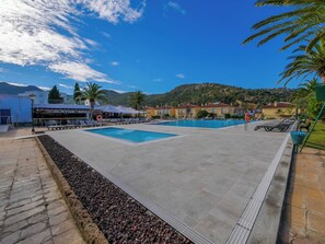 Un espace piscine aménagé avec des chaises longues et des parasols, entouré de bâtiments et de palmiers.