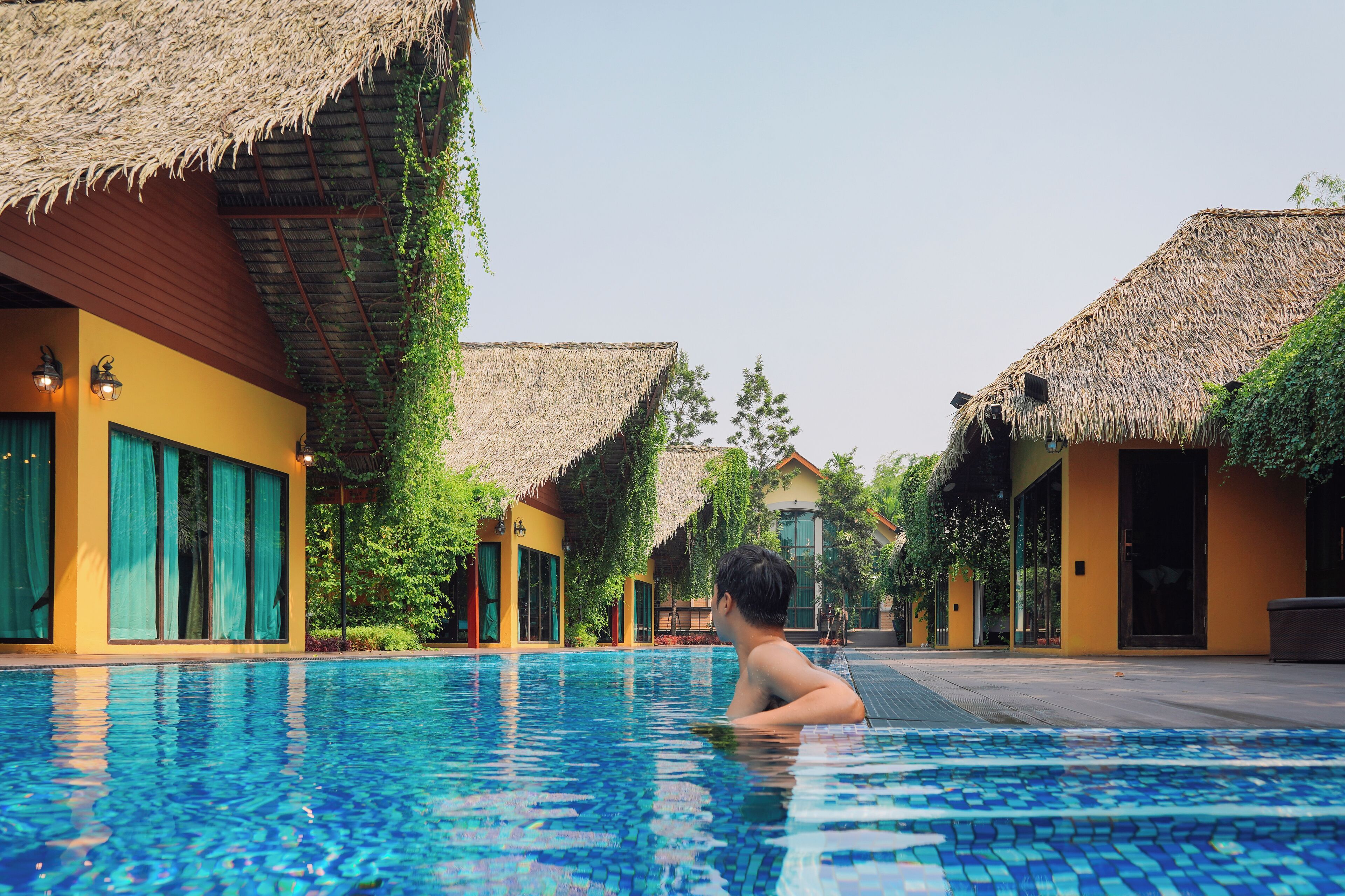 A swimming pool with a person in the water, surrounded by buildings with thatched roofs and large windows.