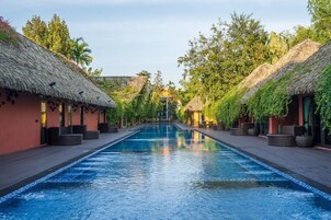 Una piscina larga con estructuras de techo de paja a ambos lados, rodeada de abundante vegetación.