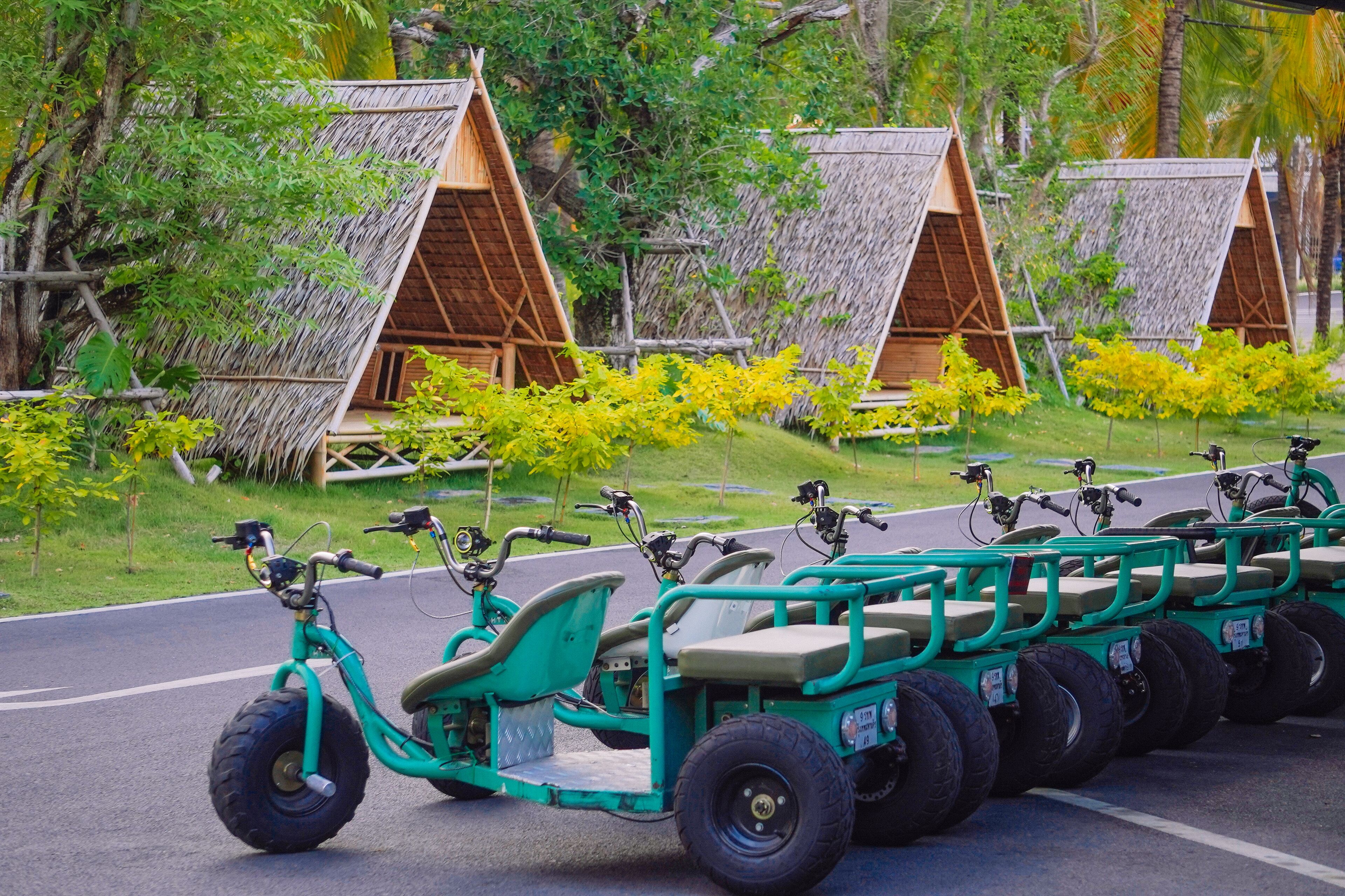 A row of green, four-wheeled, motorized carts parked on a road with thatched-roof huts in the background.