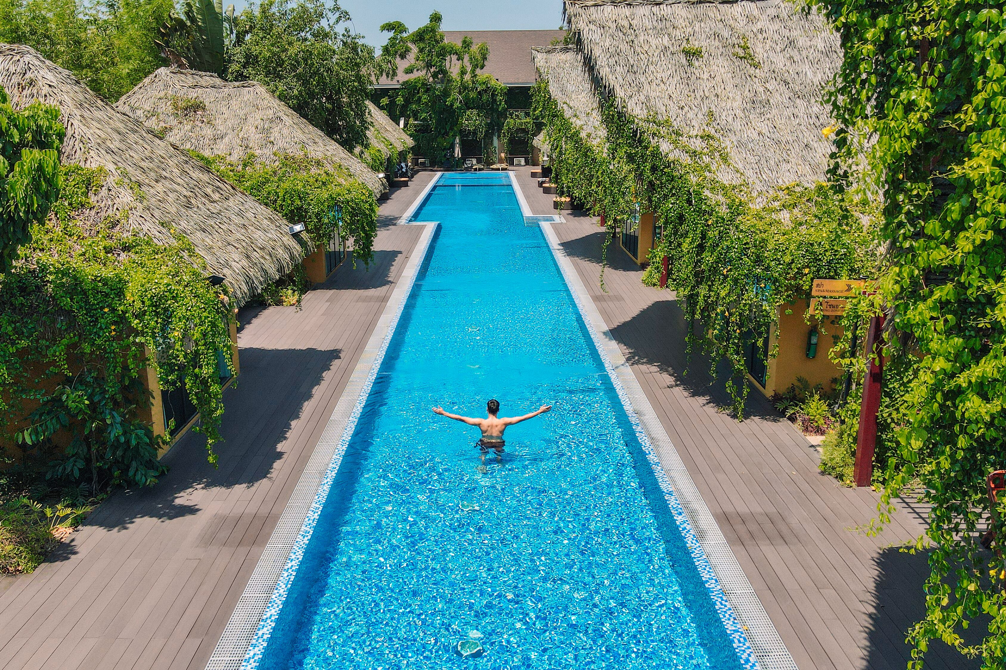 A swimming pool with a person in the water, surrounded by thatched-roof structures and greenery.