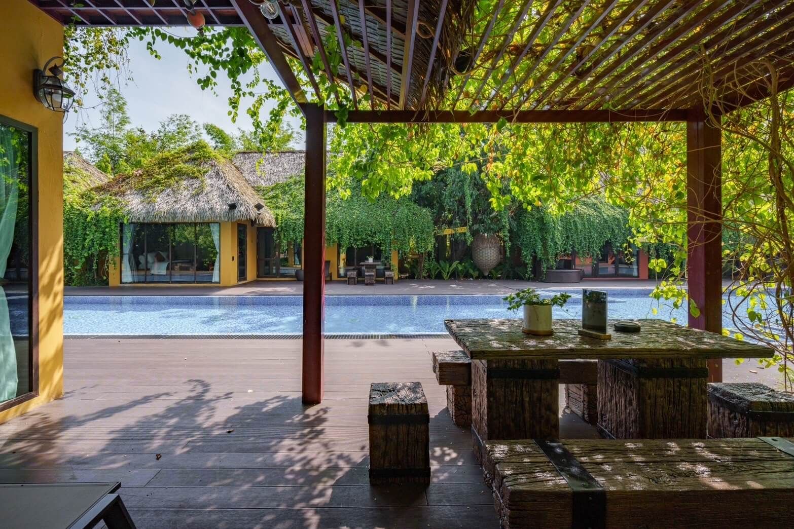 A poolside area with wooden tables and benches, a thatched-roof building, and lush greenery.