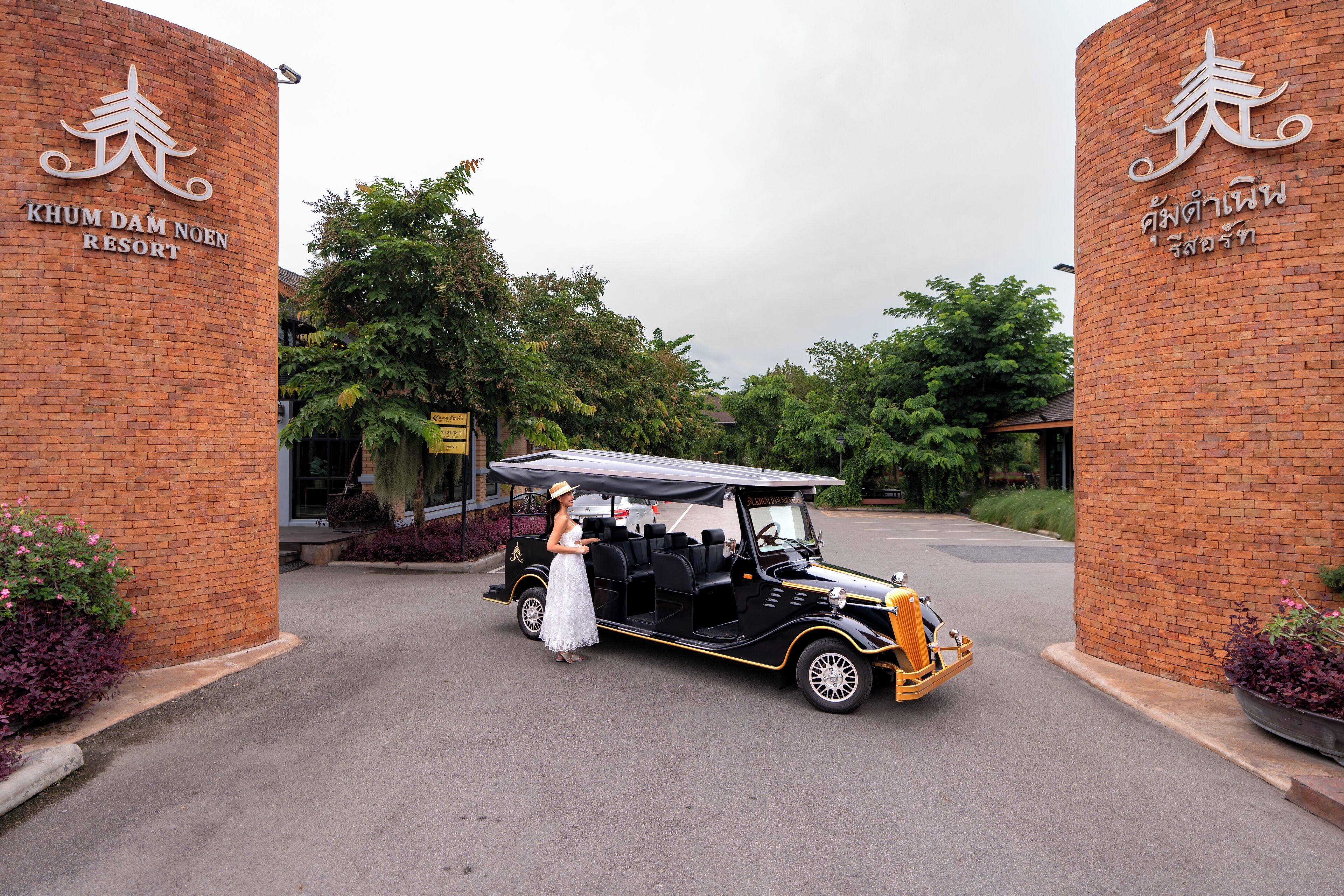 A vintage car parked at the entrance of Khum Dam Noen Resort, with a woman standing beside it.