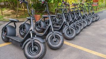Uma fileira de scooters elétricas estacionadas numa área pavimentada com linhas amarelas, rodeada por vegetação.