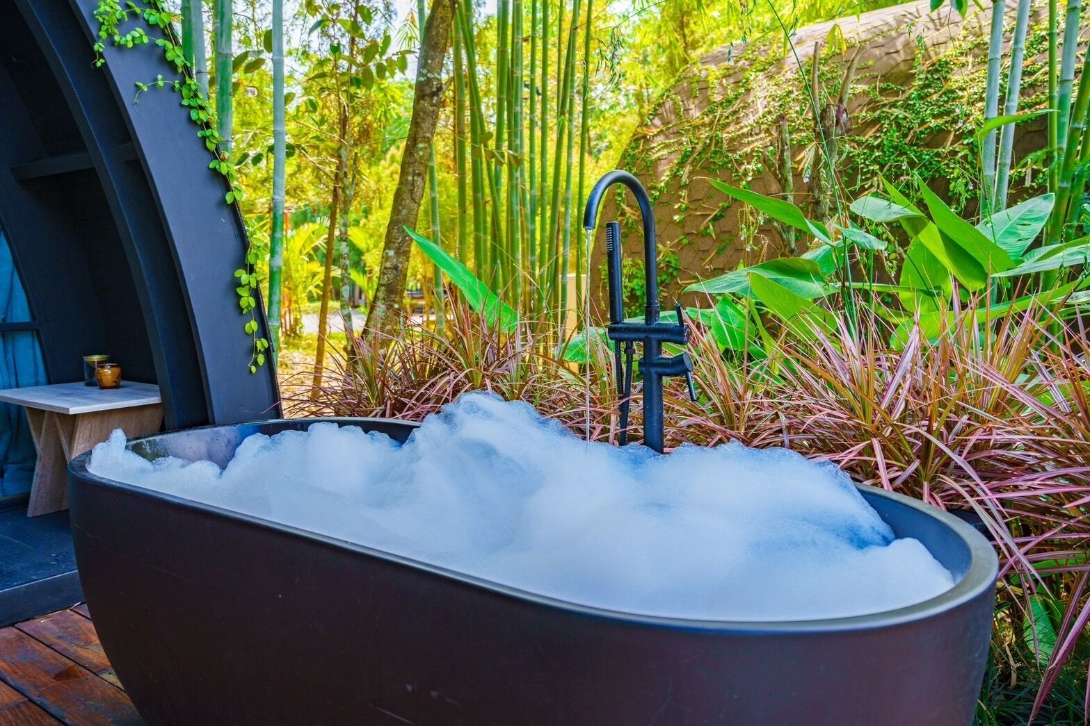 A large, dark-colored bathtub filled with ice, surrounded by lush greenery and bamboo plants.