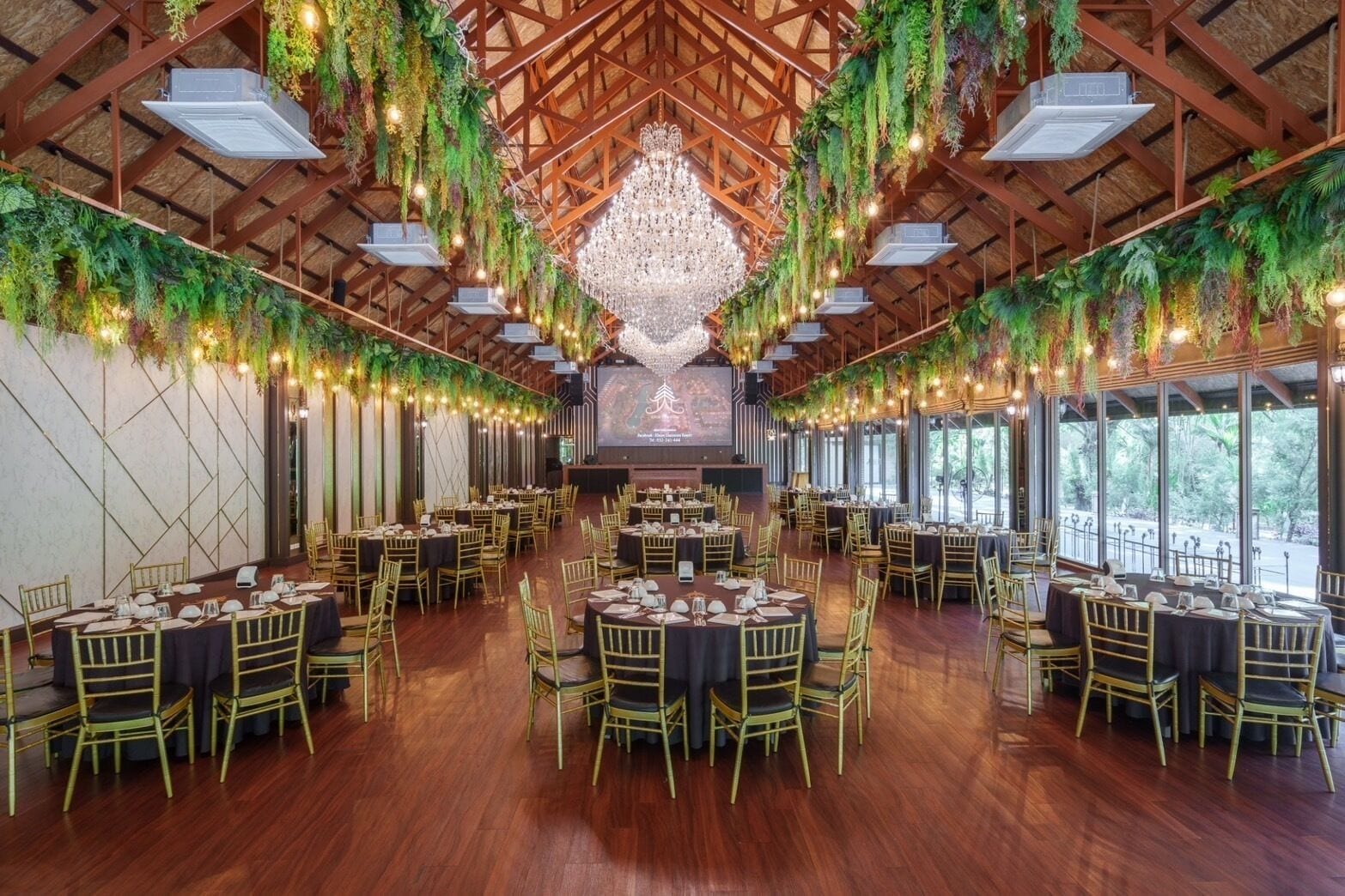 A spacious banquet hall with round tables and chairs, a large chandelier, and greenery hanging from the ceiling.
