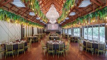 A spacious banquet hall with round tables and chairs, a large chandelier, and greenery hanging from the ceiling.